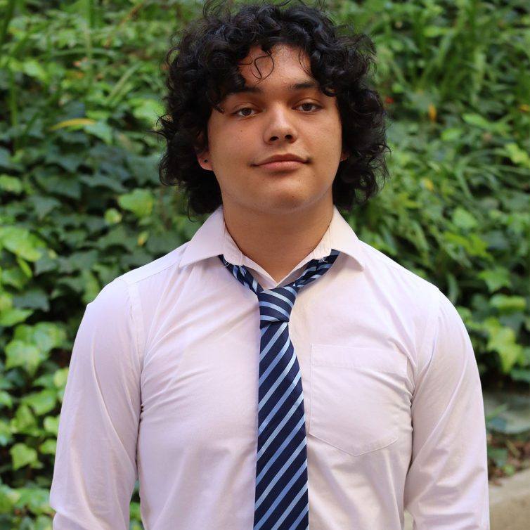 Braulio-copy Headshot of scholar. Green plants behind scholar. Scholar has shoulder length wavy black hair. They are wearing a white buttoned up shirt with a dark blue and light blue striped tie.