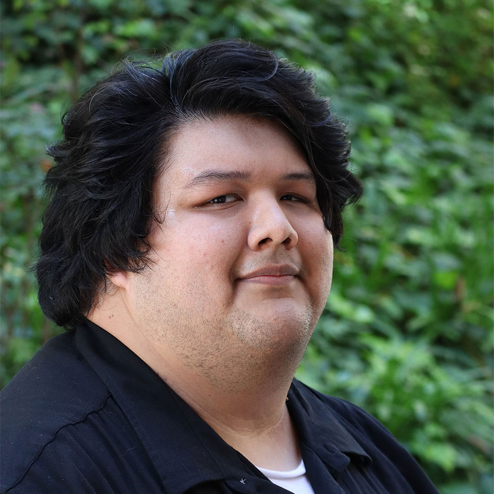 Eli-1 Headshot of scholar. Green plants behind scholar. Scholar has shoulder length wavy black hair. They are wearing a black button up shirt with a white shirt underneath. He has a closed smile with two dimples.