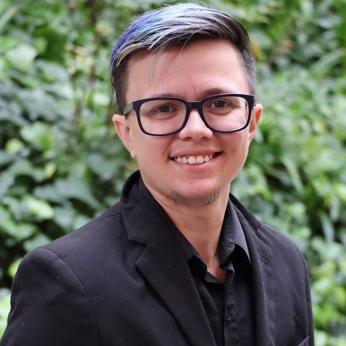 Noahnew Headshot of scholar. Green plants behind scholar. Scholar has short light purple dyed hair off to one side. They are wearing a black button up shirt inside of a black dress coat. They are wearing black round glasses.