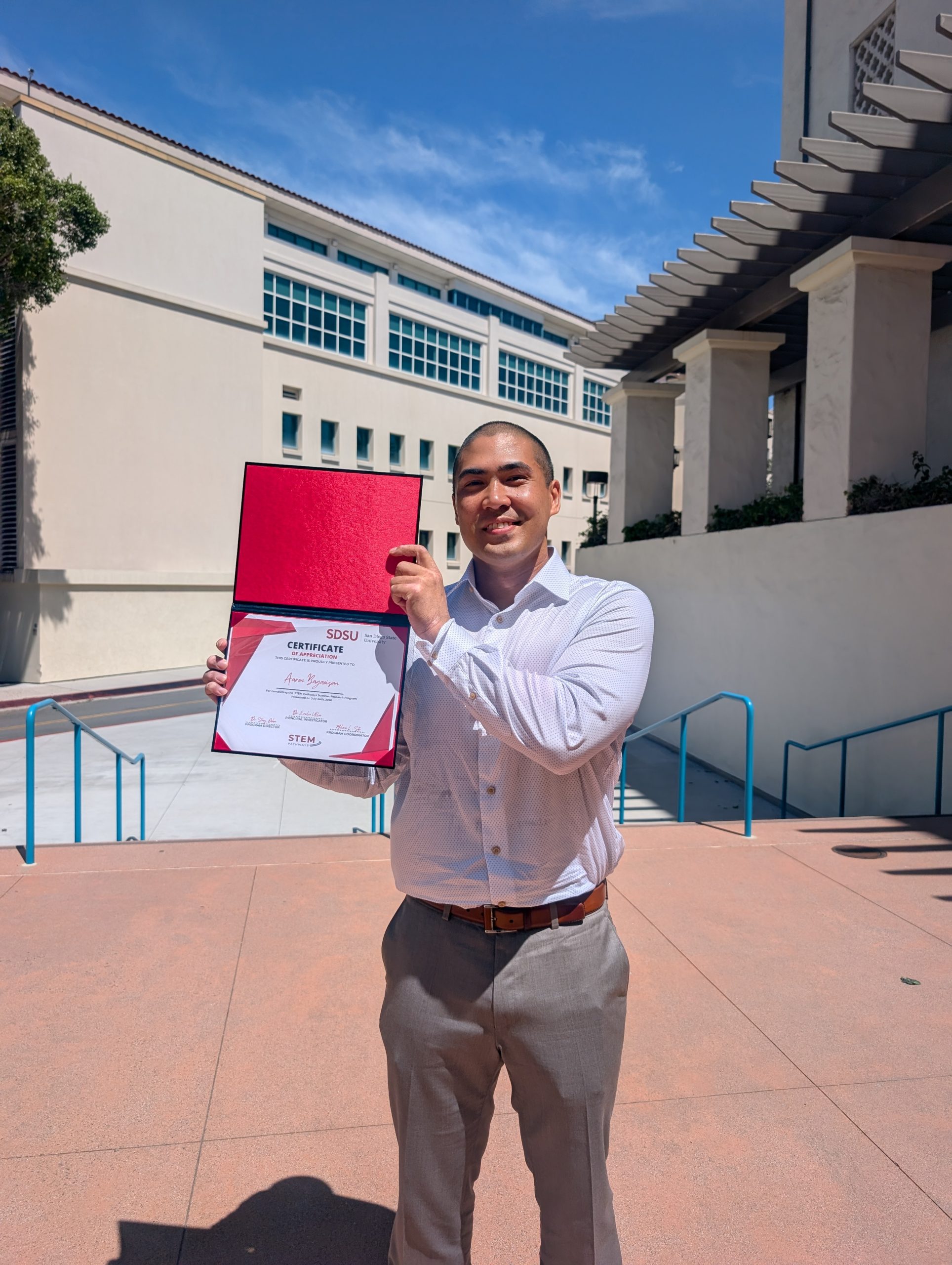 Aaron Bagaoisin is all dressed up in his white button up collar shirt and beige slacks. A brown belt holds the pants up. He stands outside showing off his certificate for the STEM Pathways Summer Research Program. We also want to congratulate him we just found out he was accepted in the MARC program. Keep it up!