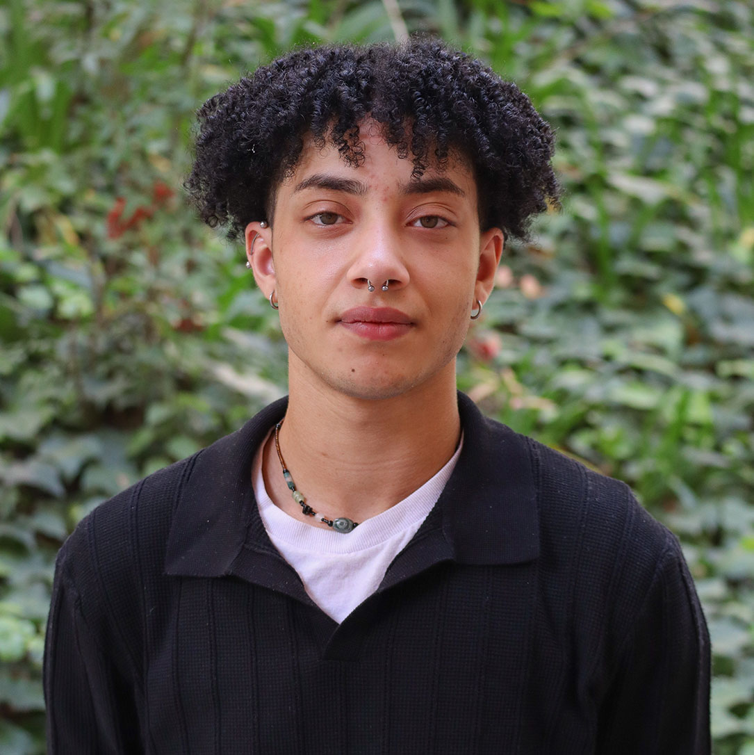 Green leaves in the background and a few red flowers. Scholar has tight curly thick black hair up to their ears. They have a nose ring and two silver hoop earrngs. They have a earring at the top of their ear cartilage a bar earring. They wear a beaded necklace. They wear a white undershirt under a black v- neck sweater.