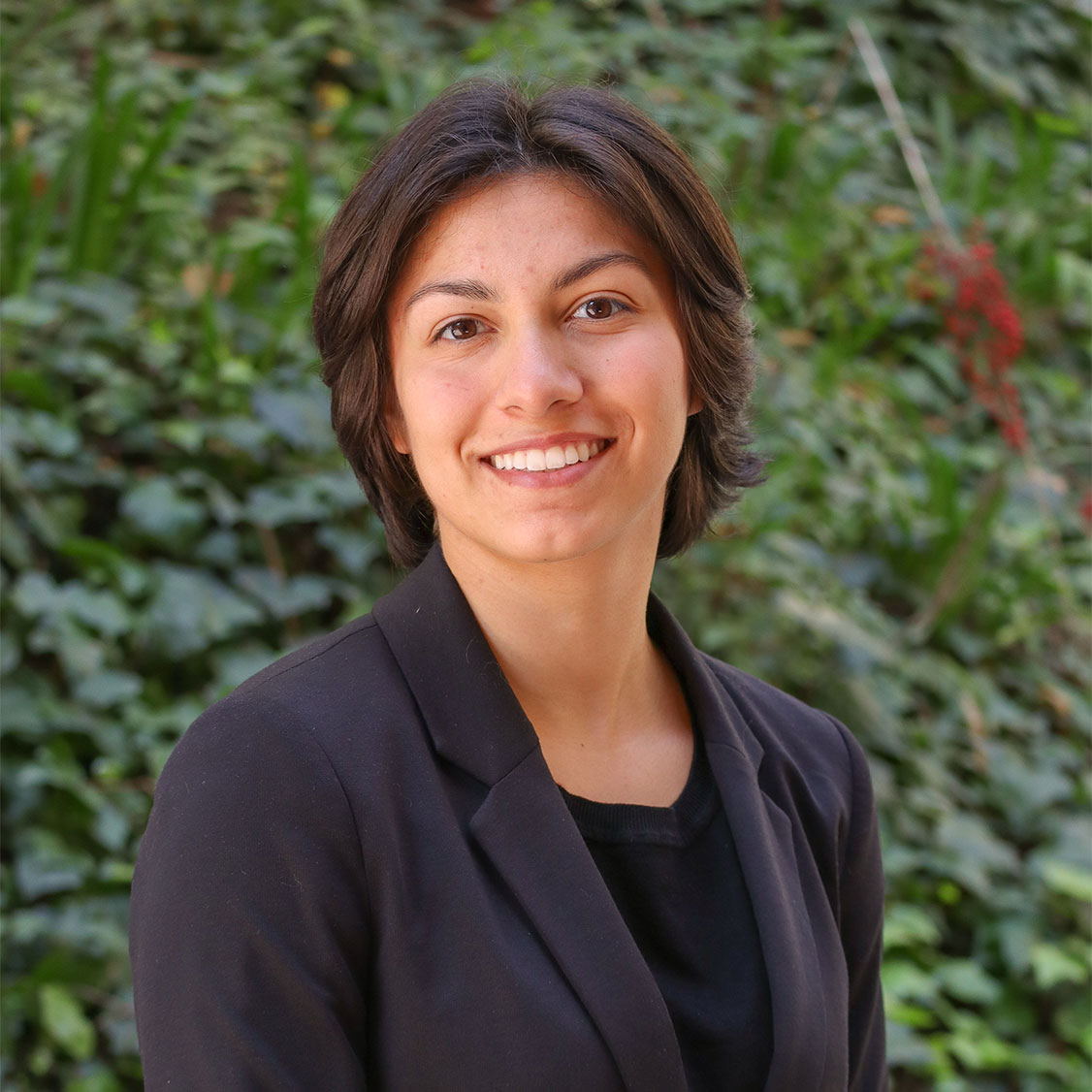 Green leaves in the background and a few red flowers. Scholar has wavy brown hair a little bit past their ears. They wear light brown lipstick. They waera black suit jacket and a black shirt underneath.
