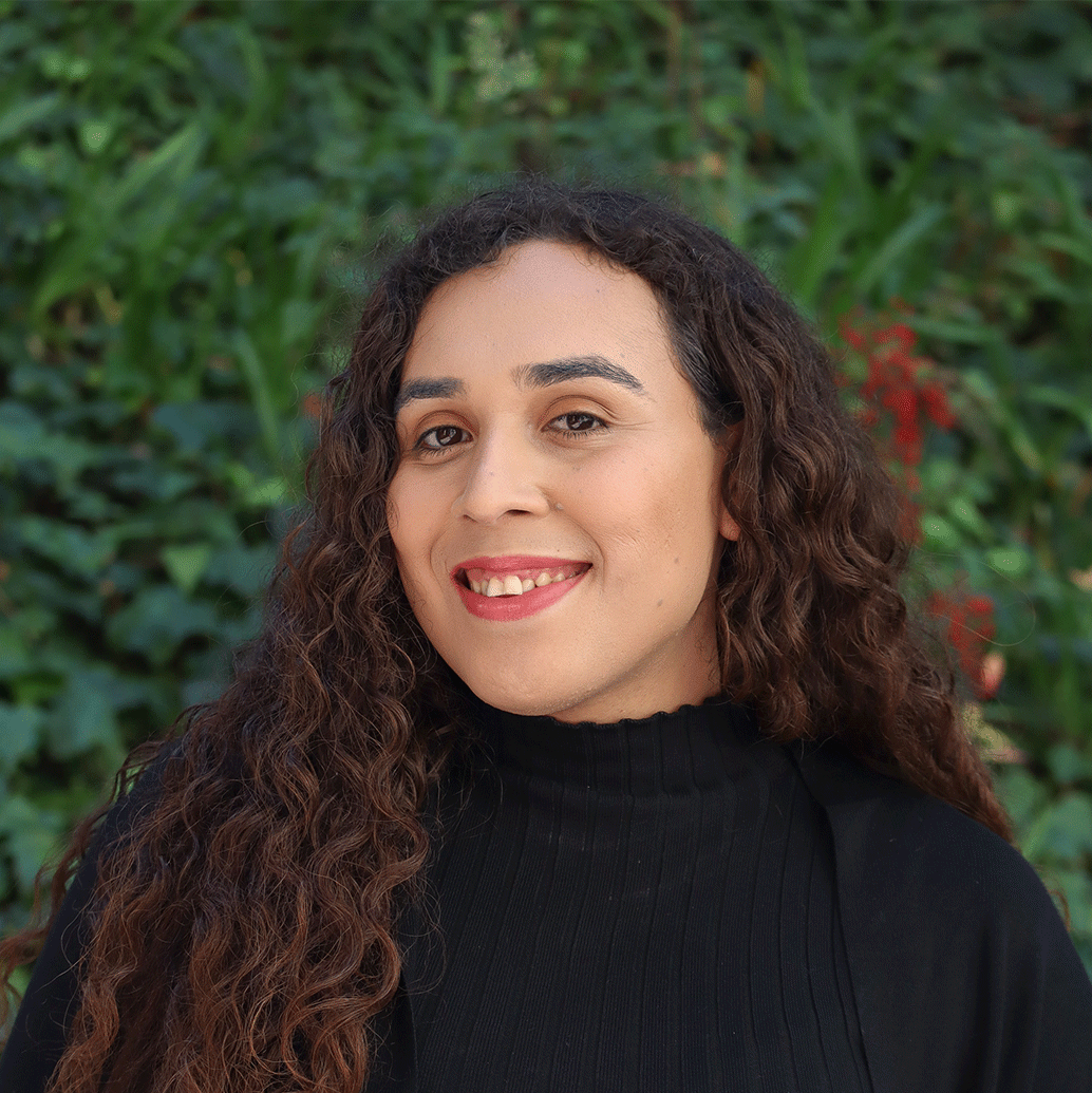 Green leaves in the background and a few red flowers. Scholar has long curly brown hair up almost up to their waist. They are wearing light pink eyeshadow and lipstick. They are wearing a black sweater.