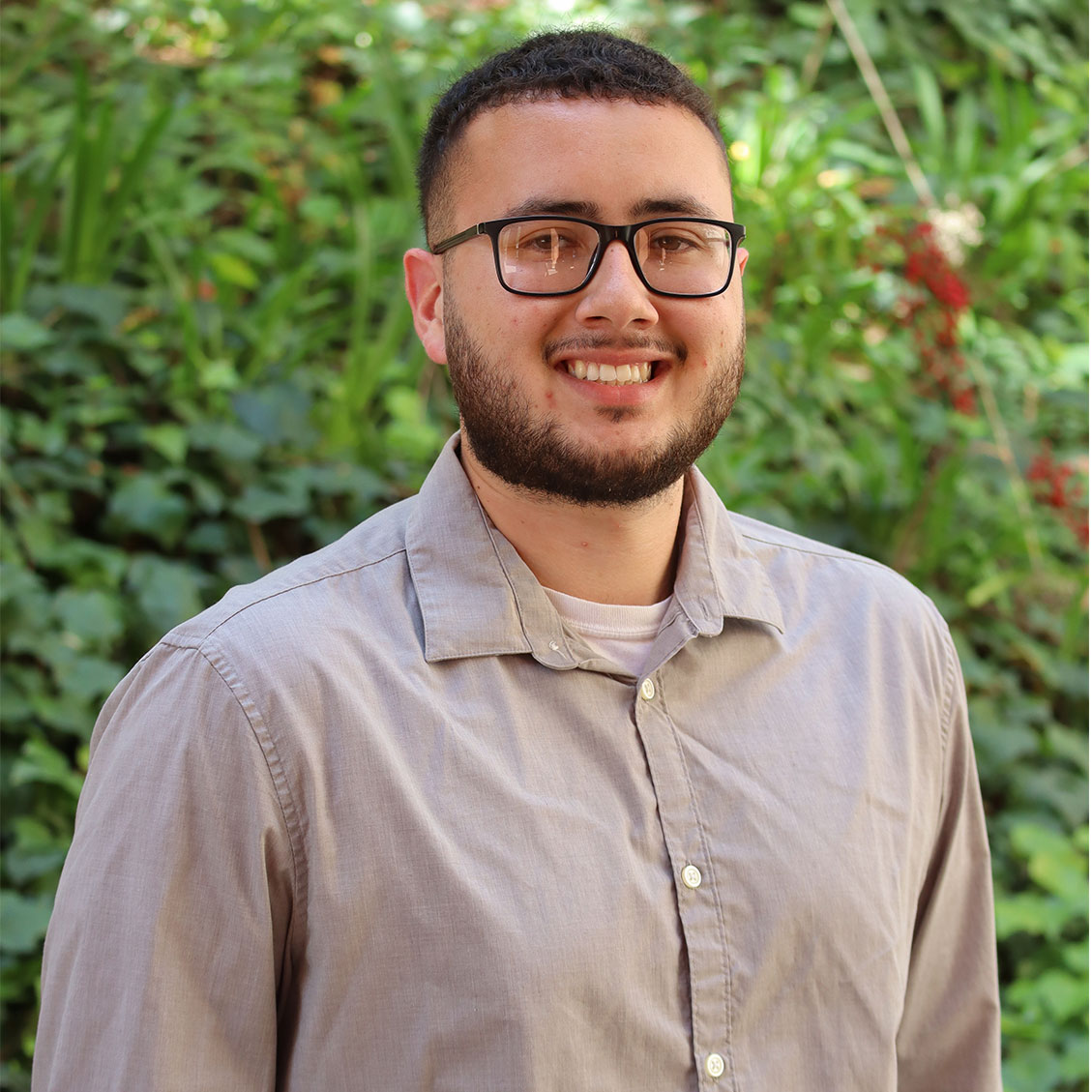 Green leaves in the background and a few red flowers. Scholar has tight curly thick black. He has a thick beard. He has a big smile. He has round square black glasses. He wears a white undershirt underneath a grey button up color dress shirt.