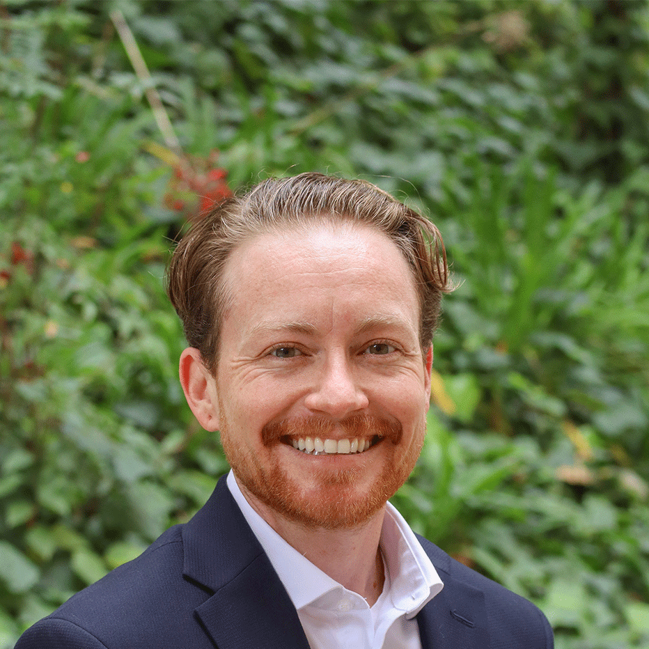 Green leaves in the background and a few red flowers. Scholar has wavy brown reddish hair up to their ears. They have a red light beard. Is wearing a black jacket coat and a white buttoned down colar shirt inside the coat.