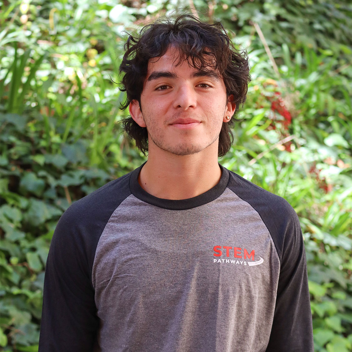 Green leaves in the background and a few red flowers. Scholar has wavy black hair up to their neck. Has very light black facial hair. They have one stud earring and is smiling with a closed mouth. They are wearing a two color grey STEM Pathwyas long sleeve shirt. STEM is written in red and Pathways is written in white with a road at teh end of it.