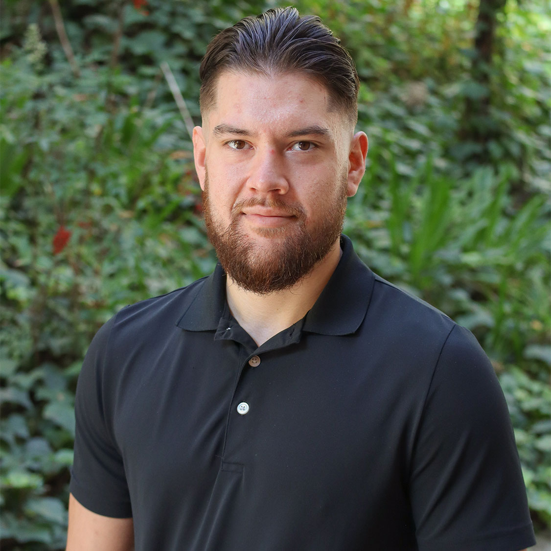 Green leaves in the background and a few red flowers. Scholar has wavy brown reddish hair up to their ears. They have reddish brown facial hair in the form of a beard.Is waering a black polo.