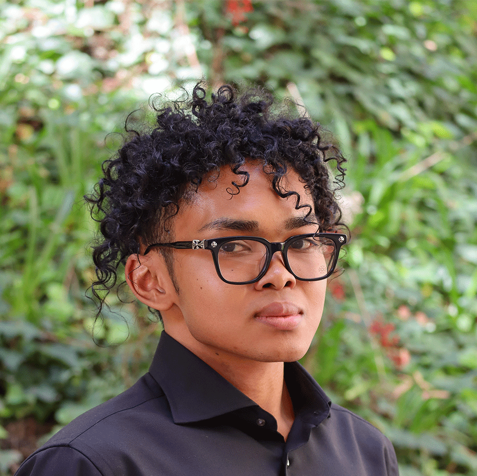 Green leaves in the background Scholar has short curly black hair. They are wearing black glasses with silver decorations on the side. They are wearing a button down black shirt.