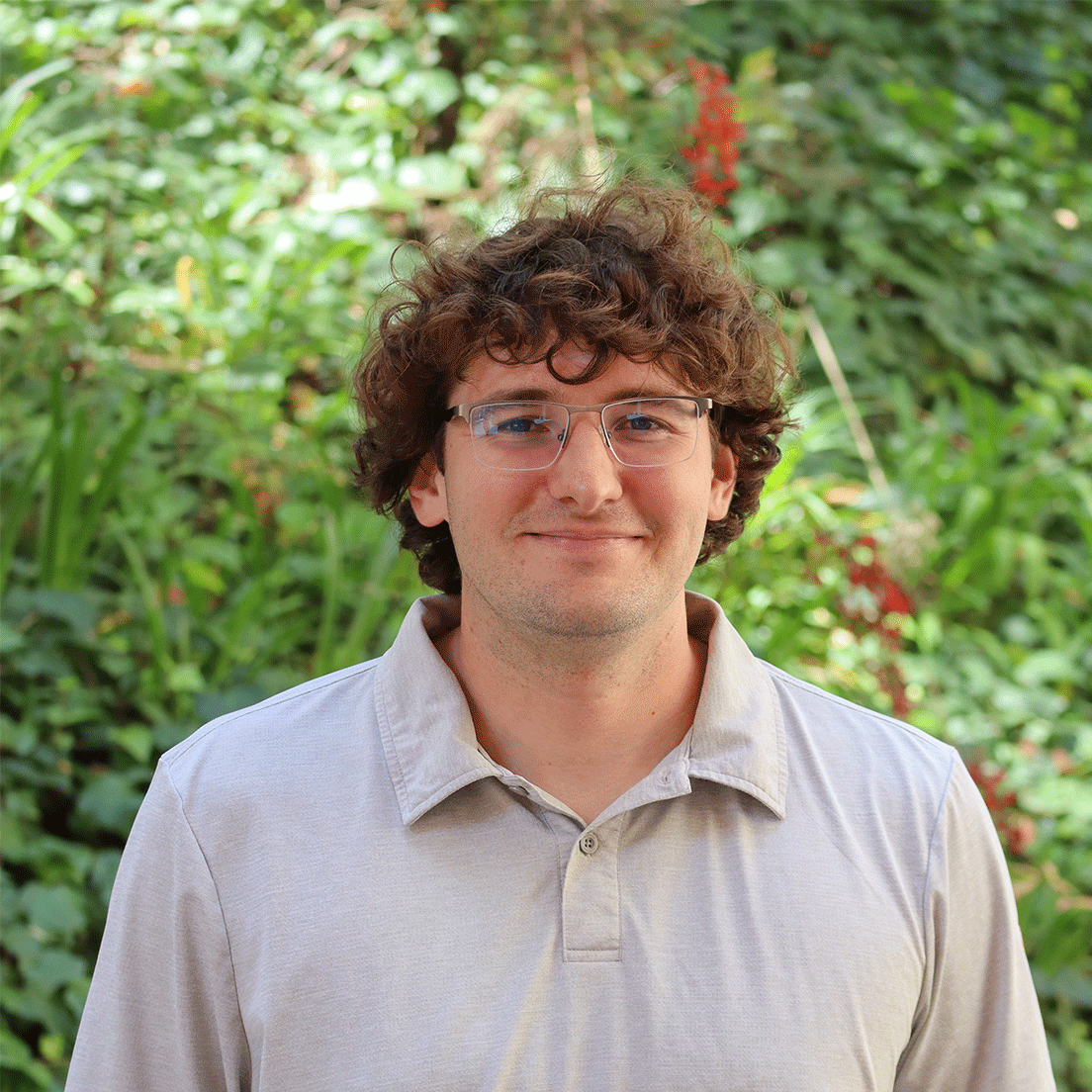 Green leaves in the background and a few red flowers. Scholar has curly brown hair up to their ears. They are wearing clear glasses. They are wearing a light brown polo.