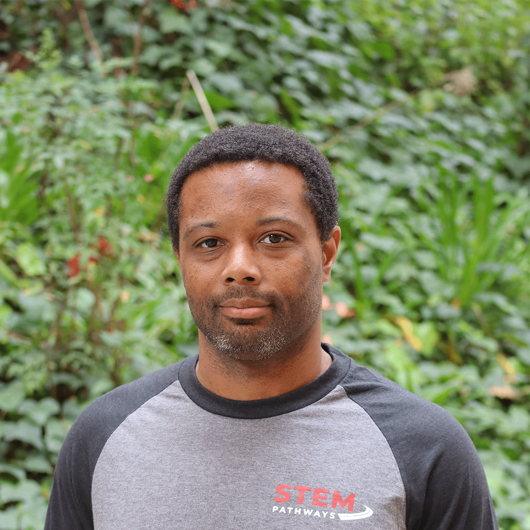Green leaves in the background and a few red flowers. Scholar has short black tight curly hair. Has a goatee with some facial hair that is a bit grey. They are wearing a long sleeve two color grey STEM Pathways shirt. STEM is written in red and Pathways is written in white with a road at the end of the word.