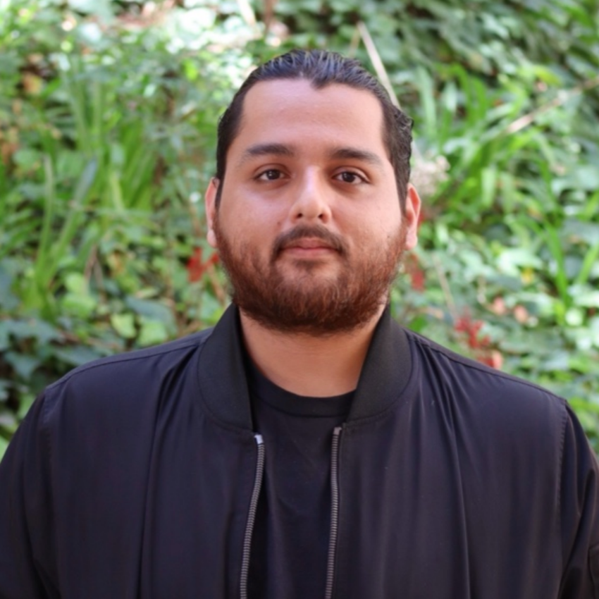 Green leaves in the background Scholar has wavy black redish hair. They have a full beard. Is wearing a black undershirt with a black opened zip up jacket .