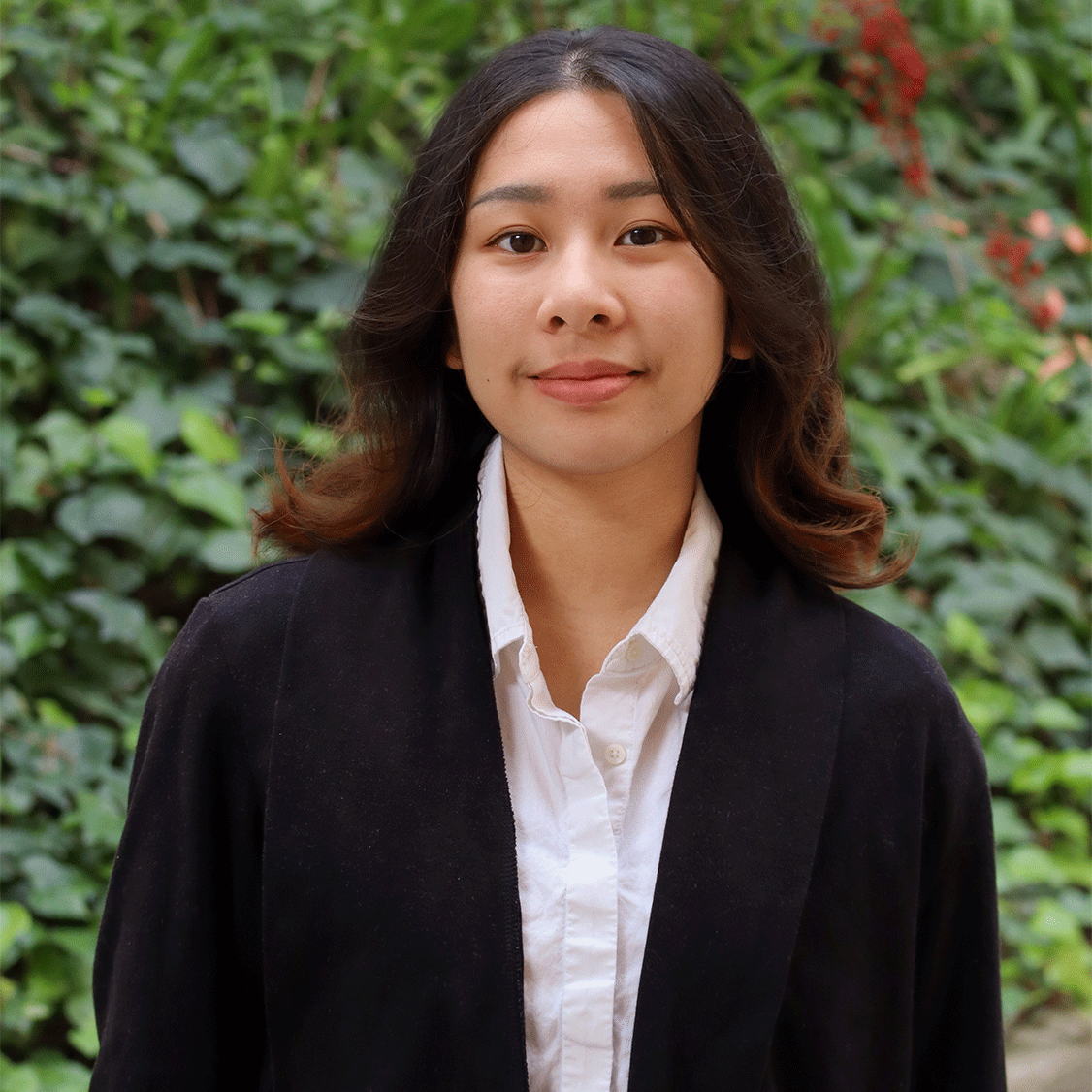 Green leaves in the background and a few red flowers. Scholar has shoulder length brown hair. The hair is curled and flipped out . Is wearing light brown lipstick. Has a black coat and a white button down collar shirt inside the black coat.