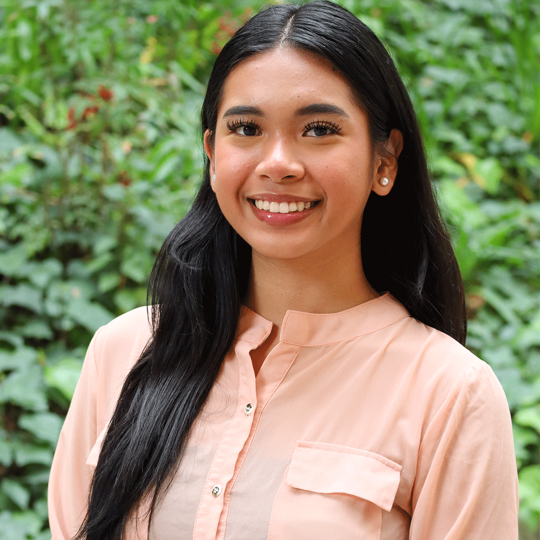 Green leaves in the background and a few red flowers. Scholar has long straight black hair. They have long eyelashes light blush and pink lipstick with lipgloss. They gave a big smile and is wearing pearl earrings. They wear a pink sheer button down shirt with a black undershirt underneath.